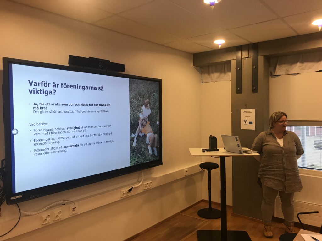 En kvinna står bredvid ett talarstativ i ett rum och håller ett föredrag. På en stor skärm visas svensk text och ett foto av två hundar. Rummet har ljusa väggar, taklampor och ett fönster med grå gardiner.