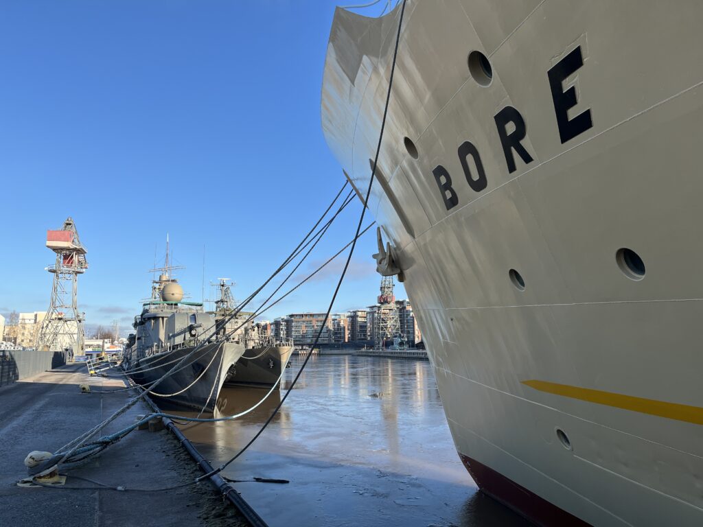 Två fartyg ligger förtöjda i en hamn; namnet BORE syns på skrovet på det stora vita fartyget i förgrunden. Ett mindre grått fartyg ligger förtöjt bredvid det. I bakgrunden syns byggnader och kranar.