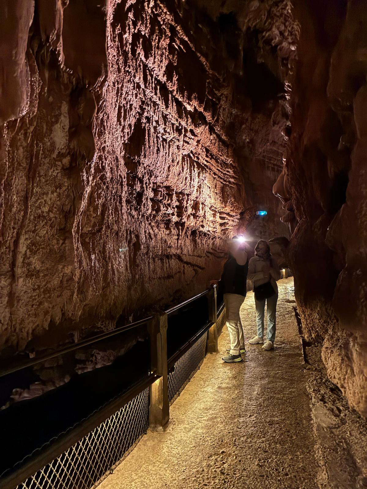 Två personer står på en upplyst gångväg inne i en grotta och tittar på den strukturerade, rödbruna klippväggen. Stigen avgränsas av ett svart räcke och nät. Grottans funktioner är upplysta av artificiella lampor.