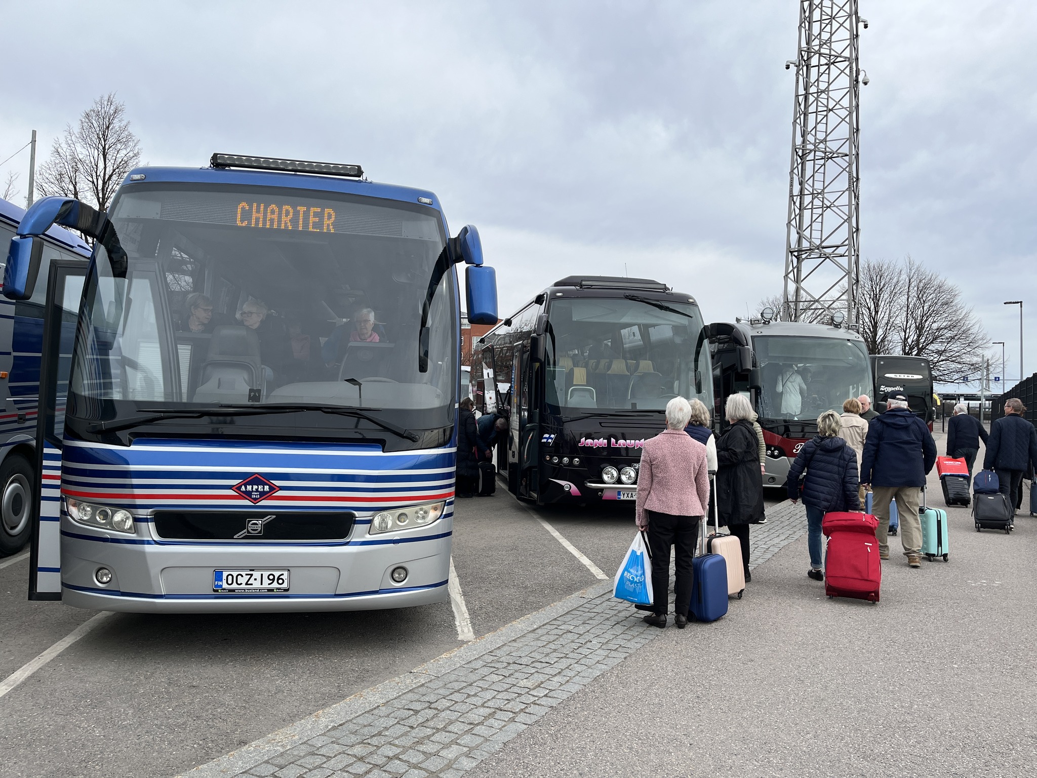 Människor med resväskor väntar på att få gå ombord på tre charterbussar som står parkerade sida vid sida på en asfalterad parkeringsplats en molnig dag. Några passagerare sitter redan inne i bussarna.