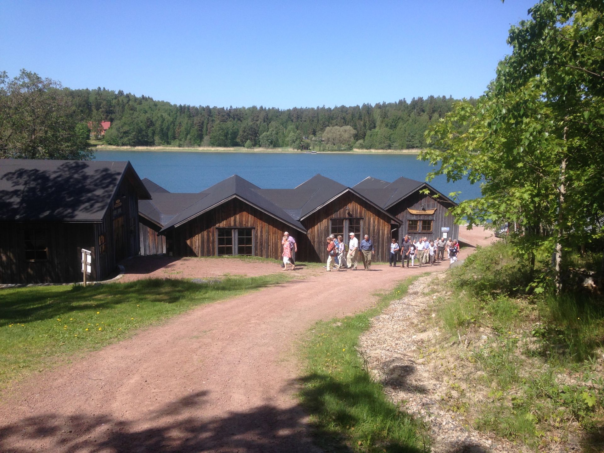 En grupp människor står nära flera träbyggnader med mörka tak vid en sjö, omgiven av träd och grönska under en klarblå himmel.