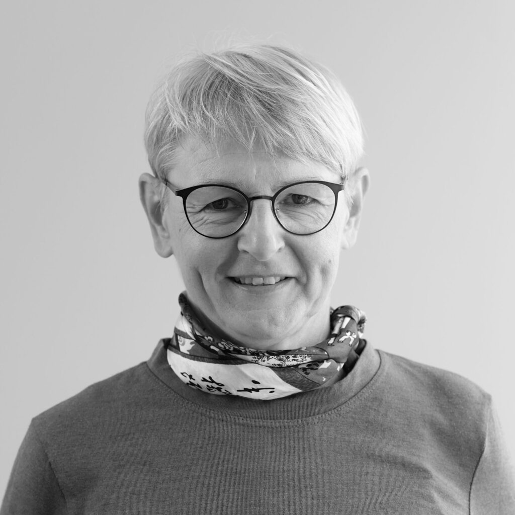 En person med kort, ljust hår och glasögon ler. Hen bär en mönstrad halsduk och en enfärgad långärmad skjorta. Fotot är svartvitt och har en enfärgad bakgrund.