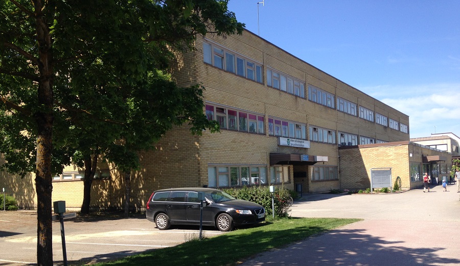 En trevåningsbyggnad i gult tegel med många fönster, omgiven av träd och gräs. En svart bil står parkerad på framsidan och i bakgrunden syns några människor promenera under en klarblå himmel.
