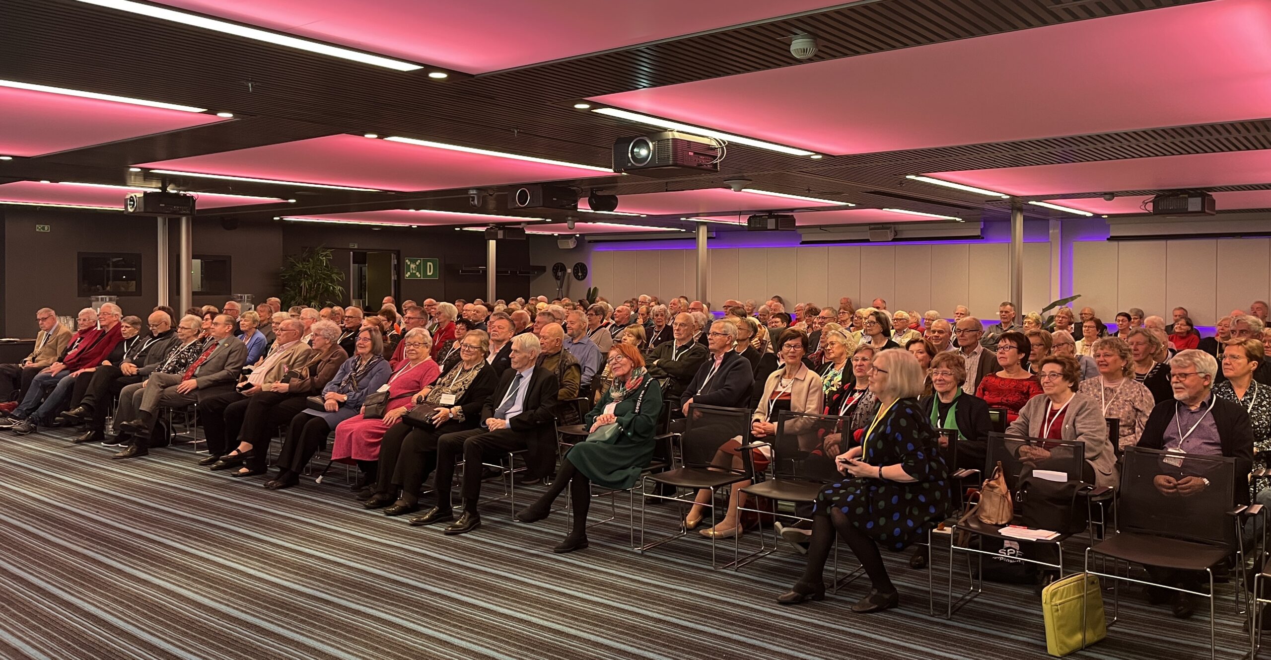 I ett modernt konferensrum med rosa taklampor sitter en stor publik, mestadels äldre vuxna, i rader av stolar vända framåt. De flesta sitter ner och lyssnar uppmärksamt, några ler eller pratar.