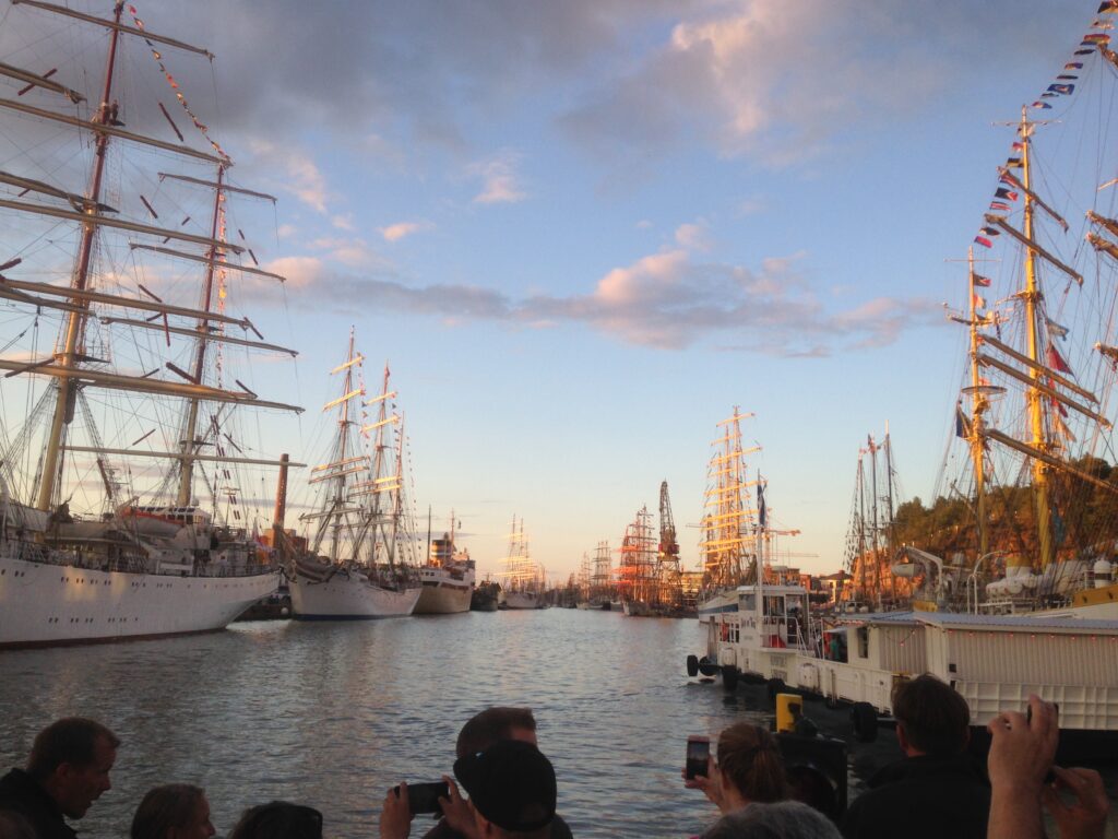 Stora fartyg med flera master och segel ligger vid kaj längs båda sidor av en hamn i solnedgången. Människor i förgrunden tar bilder av fartygen och himlen är delvis molnig med varmt ljus.