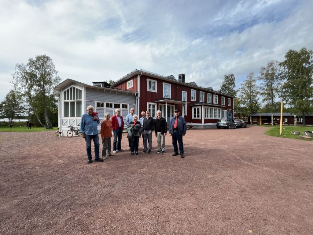 En grupp på elva personer står på en grusuppfart framför en stor röd träbyggnad med vita lister; i bakgrunden syns flera bilar och träd under en delvis molnig himmel.