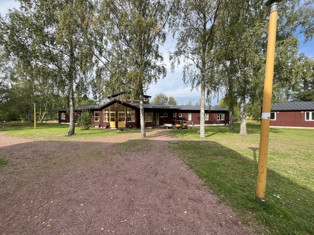 En brun träbyggnad med en täckt veranda omges av grönt gräs och höga träd. En grusgång leder fram till ingången, och i förgrunden står en gul stolpe. Himlen är delvis molnig.