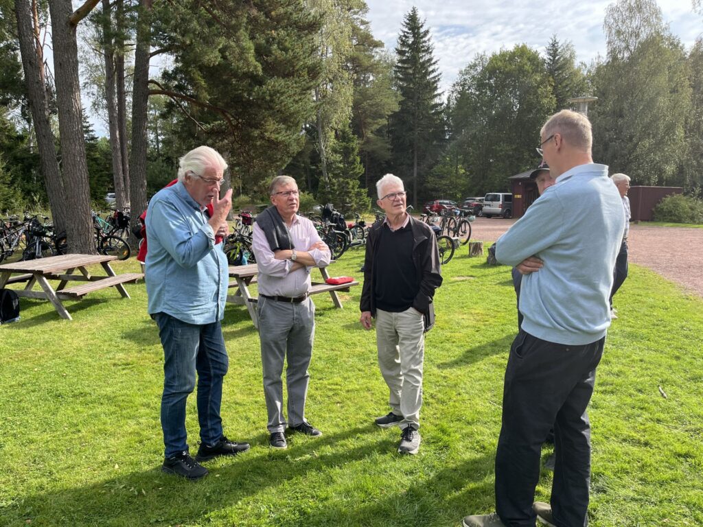 Fem äldre män står på en gräsplan och pratar nära picknickborden, med cyklar och träd i bakgrunden, en delvis molnig dag.