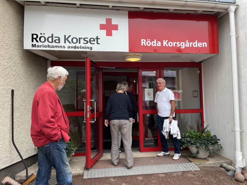 Tre personer står utanför ingången till en byggnad med skyltar där det står ”Röda Korset Mariehamns avdelning” och ”Röda Korsgården”. Två personer går in i byggnaden medan en står åt sidan.
