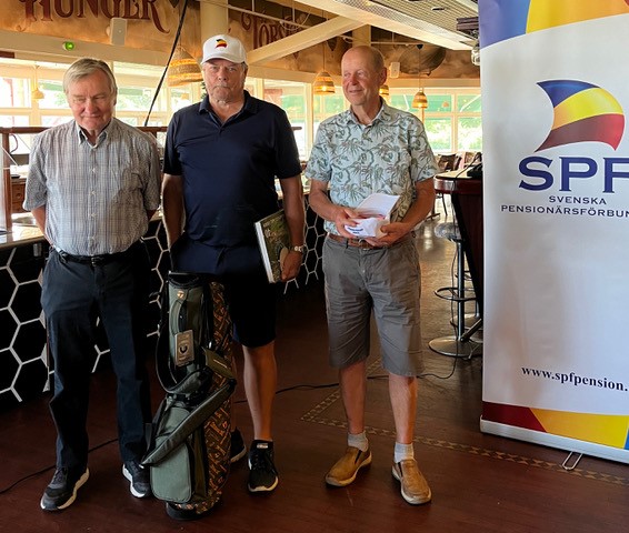 Tre äldre män står inomhus bredvid en golfväska och en banderoll från SPF Seniorerna. Två av dem håller i böcker eller mappar. De är klädda i vardagskläder och poserar för ett foto, med en inredning i restaurangstil i bakgrunden.