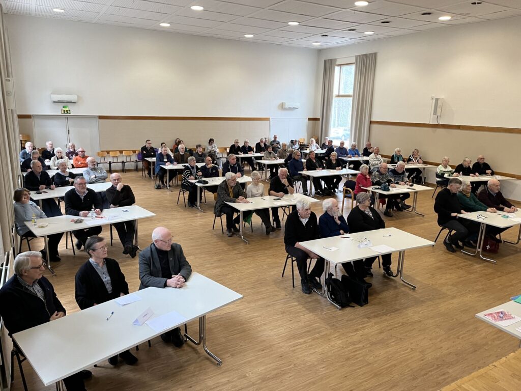 En grupp äldre personer sitter vid bord i ett stort, ljust rum och blickar rakt fram. Borden är uppställda i rader, och många av deltagarna har papper och pennor framför sig.