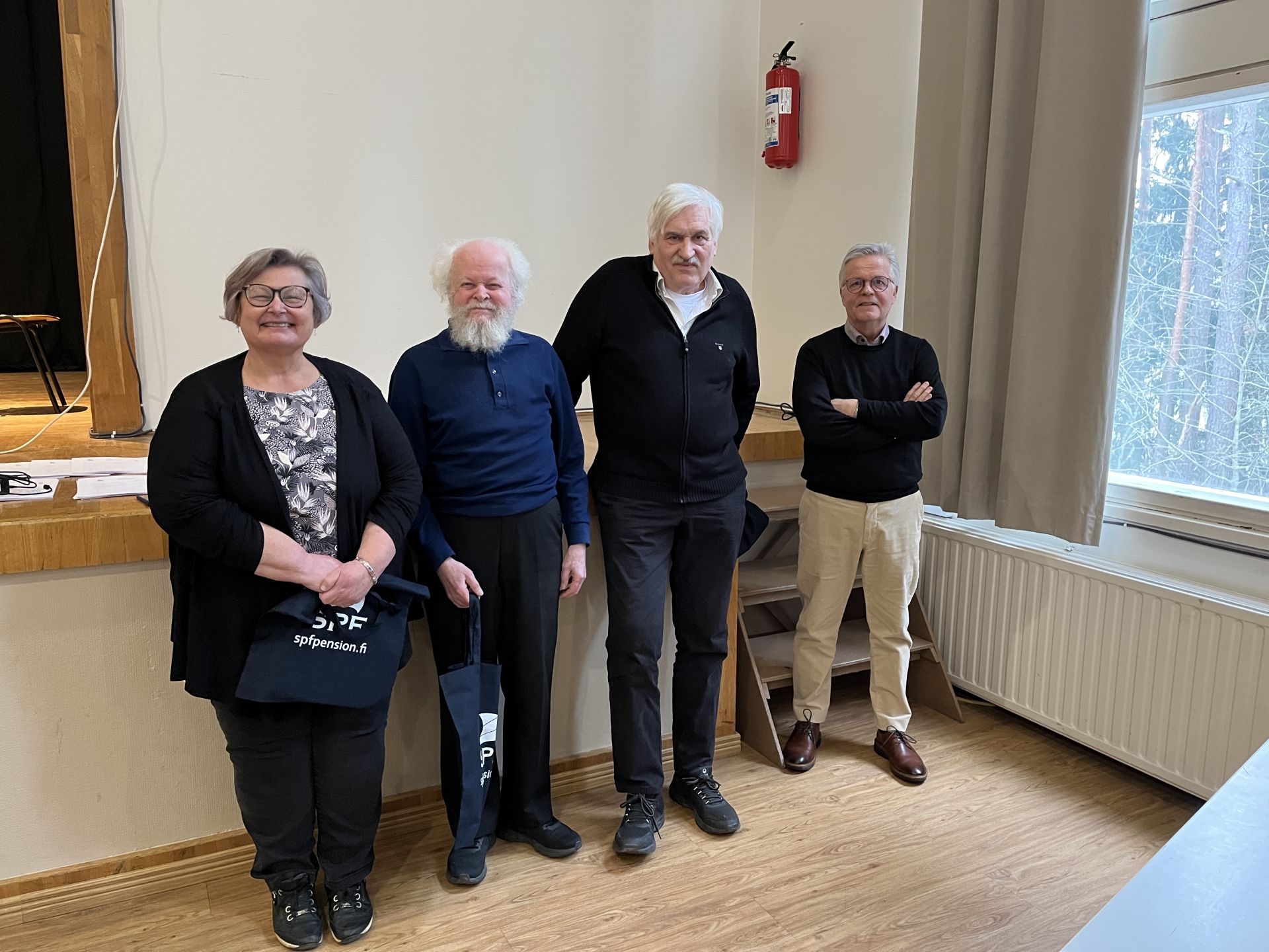 Fyra äldre personer står inomhus nära en scen och ett fönster. Tre män och en kvinna vänder sig mot kameran; två av männen har vitt hår och skägg. Rummet har trägolv, vita väggar och en brandsläckare på väggen.