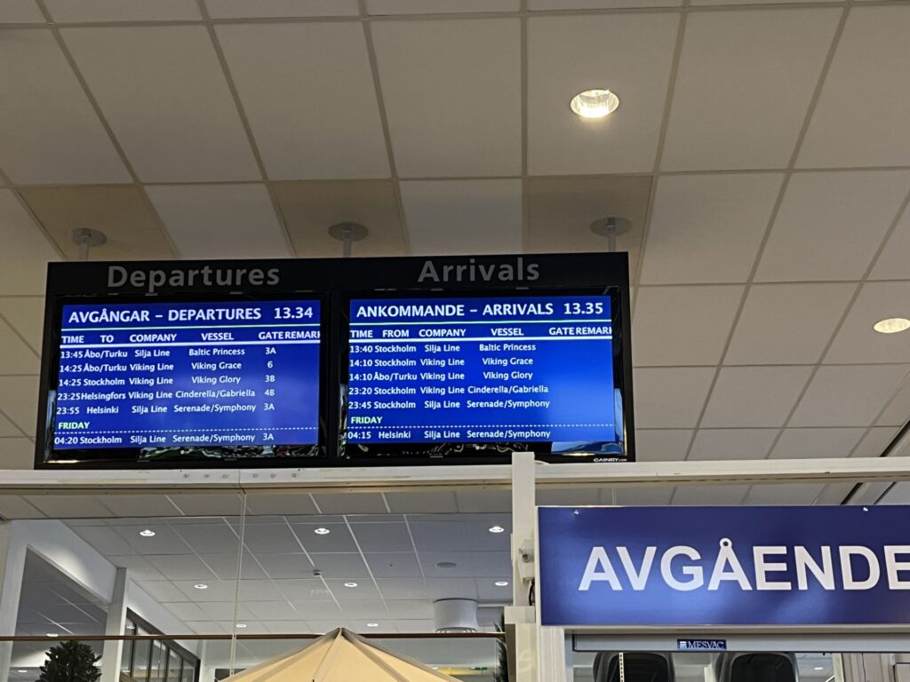 Två digitala skyltar visar färjornas avgångar och ankomster vid en terminal. Den vänstra skylten visar avgångstider, rederinamn och fartyg. Den högra skylten visar motsvarande ankomstinformation. Längst ner syns en blå skylt med texten ”AVGÅENDE”.