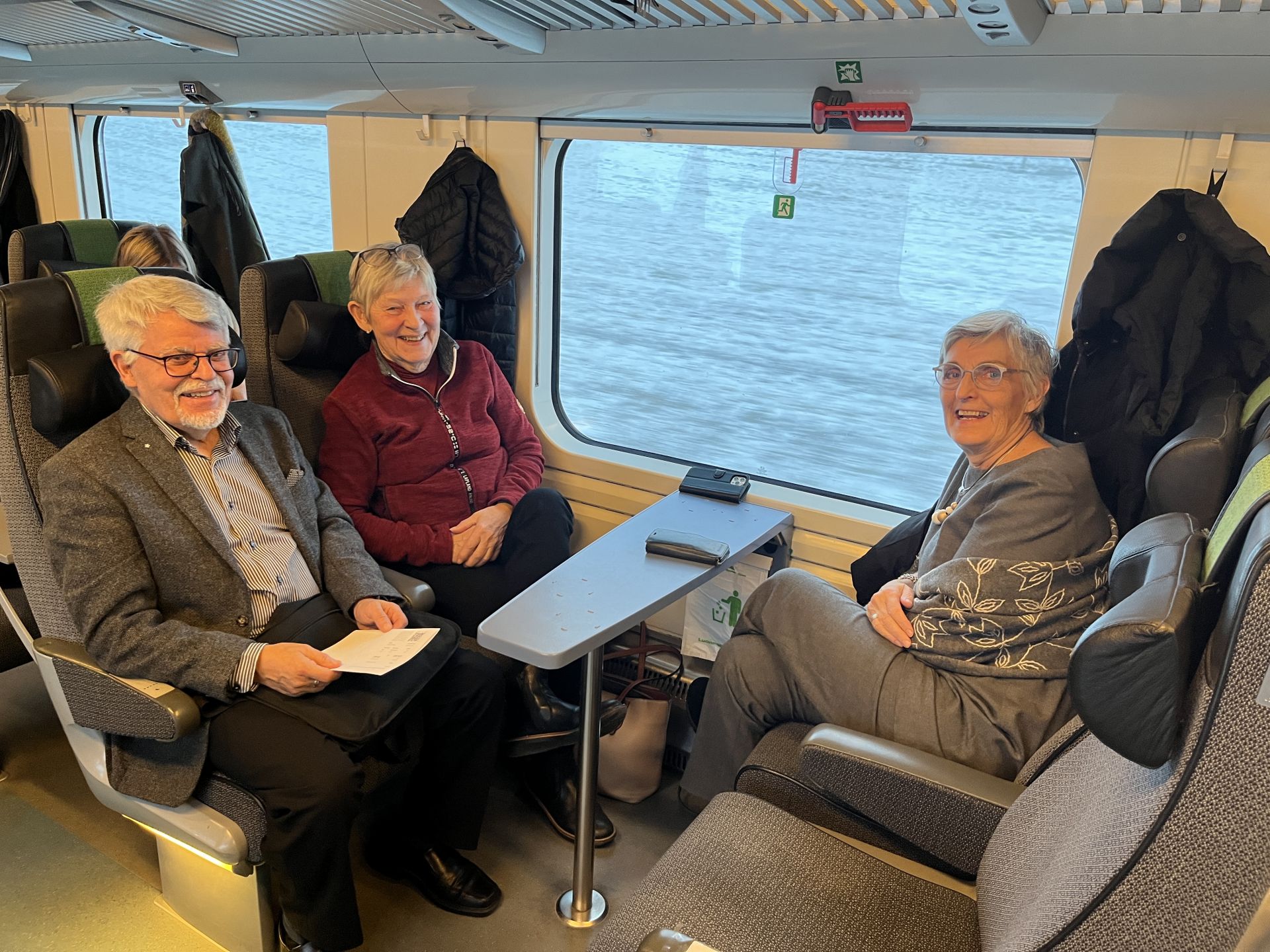 Tre äldre personer sitter tillsammans runt ett bord på ett tåg och ler mot kameran. Genom fönstret syns en suddig utsikt, vilket tyder på att tåget är i rörelse. Bakom dem hänger rockar på krokar.