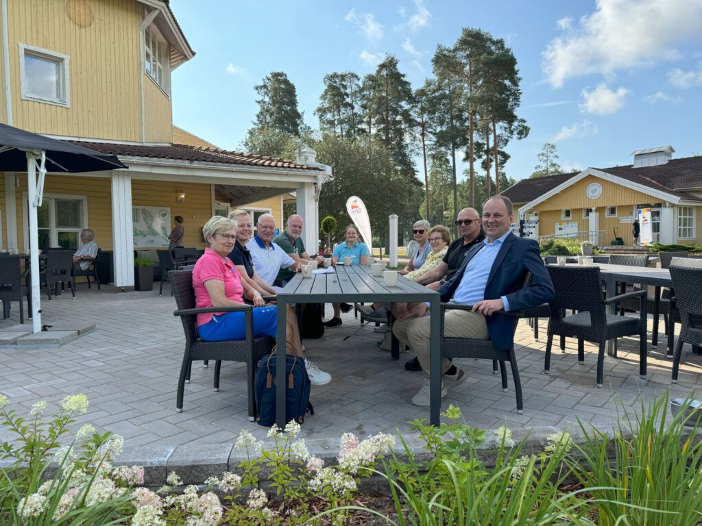 En grupp på tio äldre personer sitter runt ett rektangulärt utomhusbord på en uteplats framför en gul byggnad, med träd och blå himmel i bakgrunden. Några ler mot kameran.