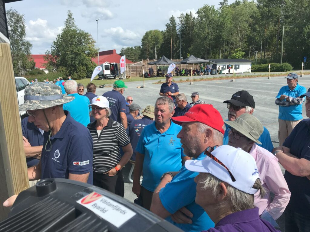 En grupp människor, främst äldre vuxna, står utomhus på en grusplan; några av dem bär hattar och solglasögon. De verkar lyssna på eller titta på något till vänster. I bakgrunden syns träd och tält.
