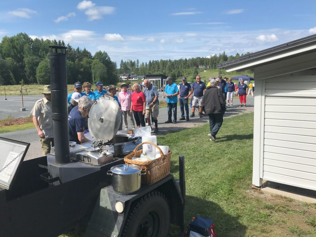En grupp människor står i kö utomhus en solig dag och väntar på att få mat serverad från en stor svart mobil matlagningsenhet. Till höger syns en vit byggnad och i bakgrunden står träd.