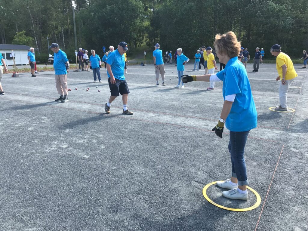 En grupp vuxna klädda i blå och gula tröjor spelar ett utomhusspel med metallbollar på en grusplan, omgiven av träd. En kvinna står i en gul ring och håller en boll, medan de andra spelarna tittar på.