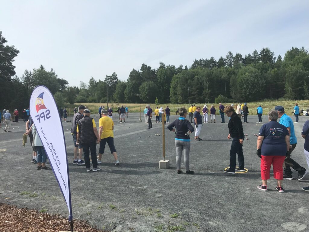 En grupp människor, varav några bär gula eller blå tröjor, har samlats på en grusplan utomhus. Området omges av träd och en SFP-flagga står i förgrunden. Flera personer spelar eller tittar på en match.