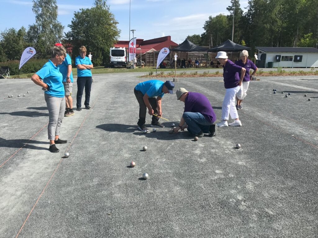 Flera personer spelar pétanque på en grusplan utomhus. Två män hukar sig ner för att mäta avståndet mellan klotarna, medan andra står upp eller tittar på. I bakgrunden syns träd, tält och en lastbil under en solig himmel.