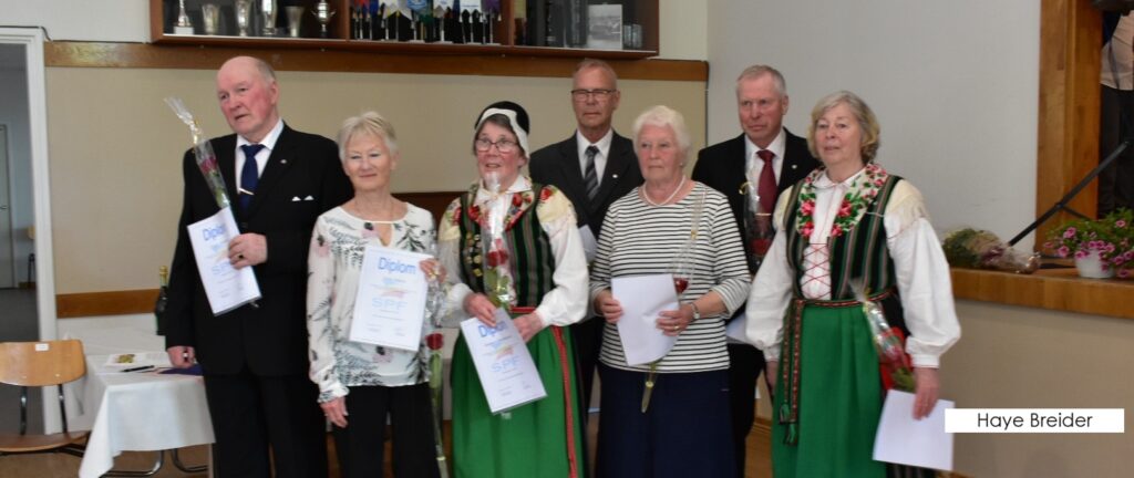 En grupp på sju äldre personer poserar inomhus. Fyra kvinnor står längst fram, varav två är klädda i traditionella folkdräkter och två i moderna kläder, och håller i examensbevis och blommor. Tre män i kostym står bakom dem.