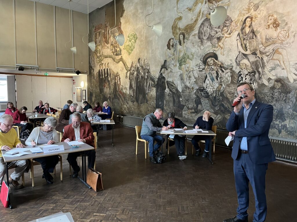 En man i kostym talar i en mikrofon längst fram i ett rum där människor sitter vid bord och skriver eller läser dokument. I rummet finns en stor väggmålning och takbelysning.