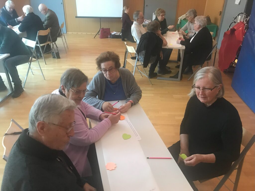 En grupp äldre sitter runt bord i ett väl upplyst rum och skriver och diskuterar med hjälp av pennor och färgat papper. Flera grupper arbetar tillsammans, och i bakgrunden syns en projektorskärm.