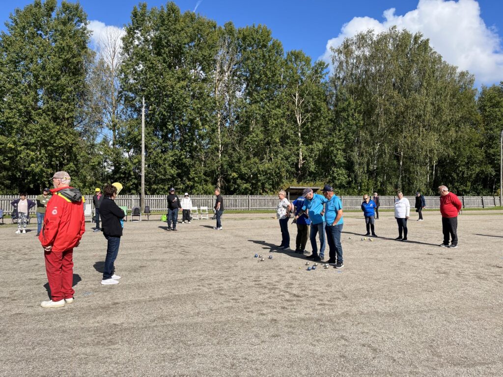 En grupp människor står i små grupper på en grusplan och spelar eller tittar på en omgång boules eller pétanque. I bakgrunden syns träd och ett trästaket under en delvis molnig himmel.