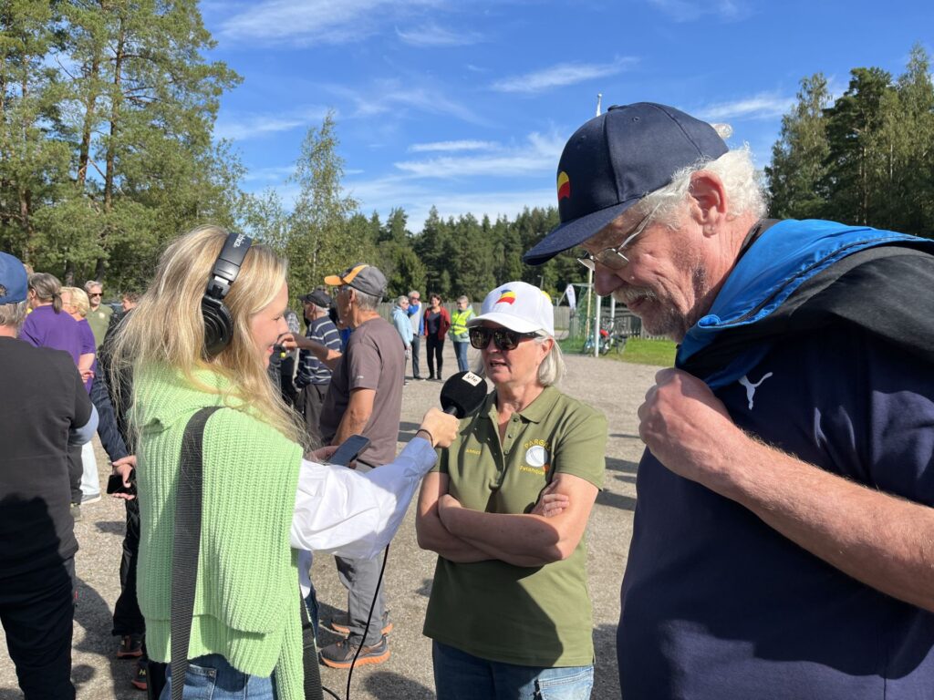 En kvinna med hörlurar på sig och en mikrofon i handen intervjuar en äldre kvinna med keps och solglasögon. En man med solglasögon står i närheten. Flera personer har samlats utomhus med träd och en blå himmel i bakgrunden.