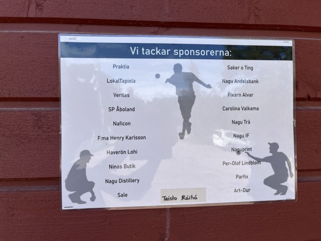 En tryckt skylt på svenska, uppsatt på en röd trävägg, visar sponsorernas namn i två kolumner, med rubriken ”Vi tackar sponsorerna”. I bakgrunden på skylten syns silhuetter av människor som spelar fotboll.