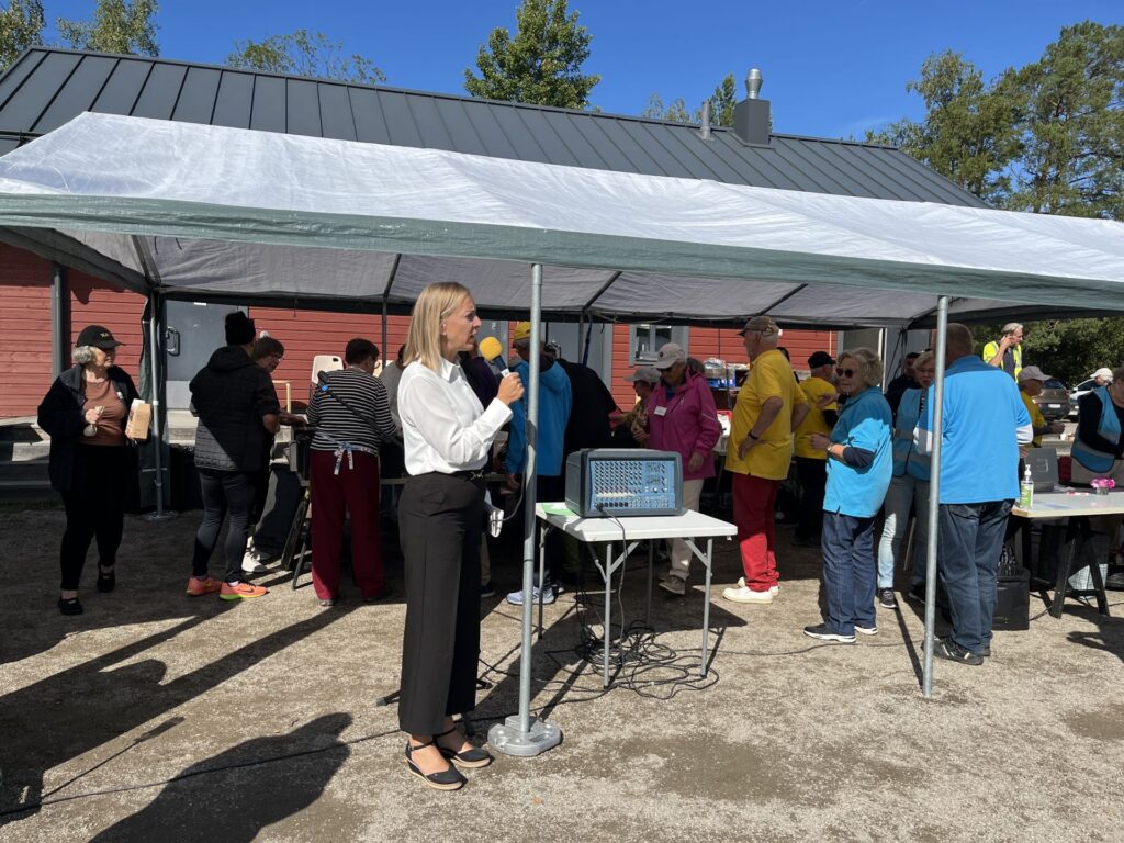 En kvinna i en vit blus talar i en mikrofon under ett tält, medan en grupp människor – varav några bär blå eller gula skjortor – samlas runt bord utomhus en solig dag.