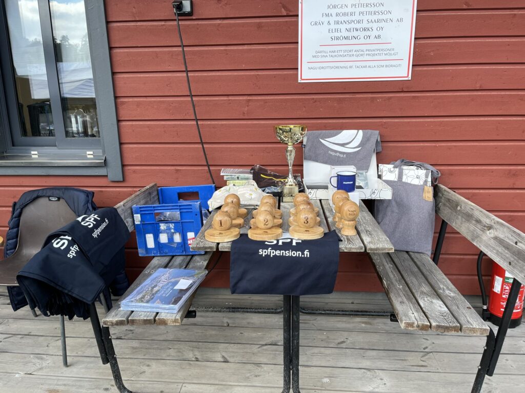 På ett träbord står flera troféer av trä, en guldpokal, mörkblå påsar med texten ”sppension.fi”, en blå låda, inslagna paket och andra föremål. Bordet står framför en röd trävägg med en skylt ovanför.