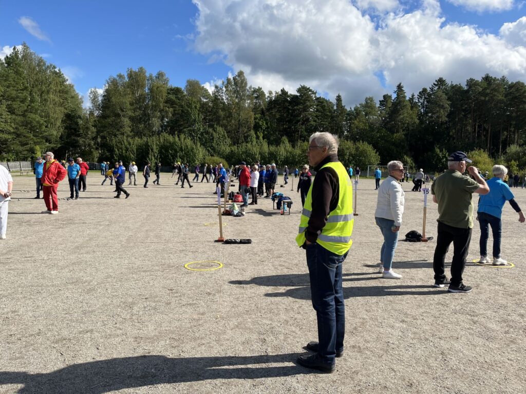 Människor har samlats på en grusplan, och några spelar ett spel med små bollar och runda markörer. En man i en gul säkerhetsväst står i förgrunden. I bakgrunden syns träd och moln.