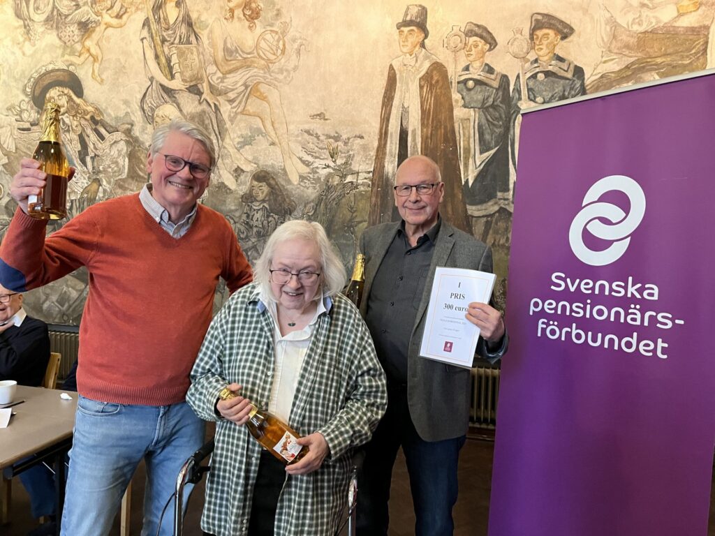 Tre äldre vuxna står inomhus, ler och håller i flaskor. En person håller i ett certifikat. Bakom dem syns en lila banderoll med Svenska pensionärsförbundets logotyp. På väggen i bakgrunden syns en muralmålning.