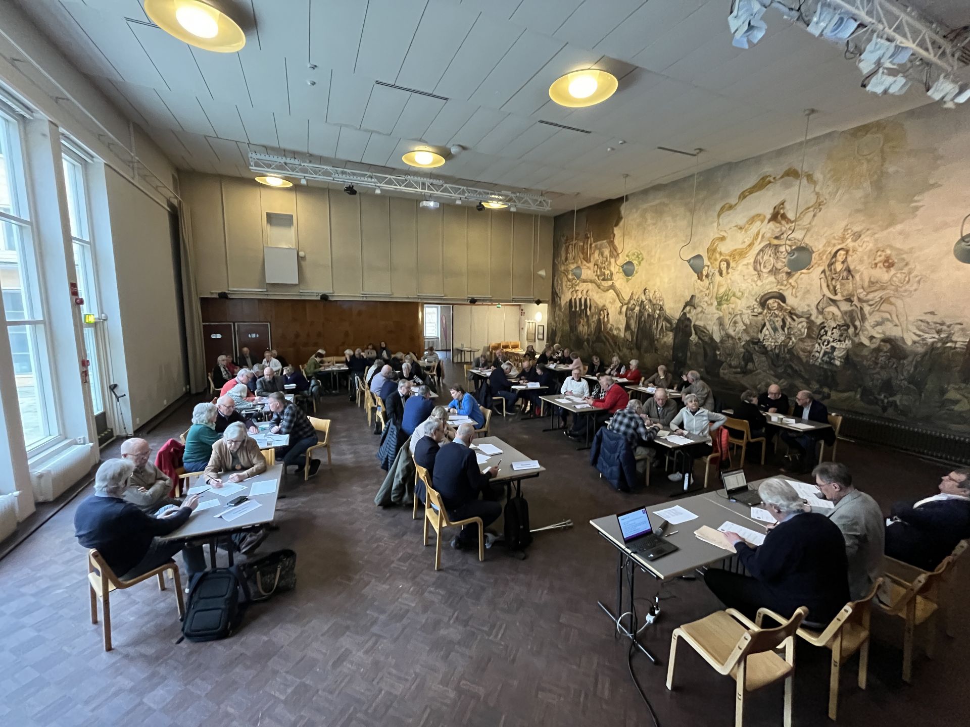 En grupp människor sitter vid bord i ett stort rum med högt i tak, stora fönster och en väggmålning på ena väggen. På flera bord ligger dokument, bärbara datorer och papper utspridda. Belysningen är stark och naturlig.
