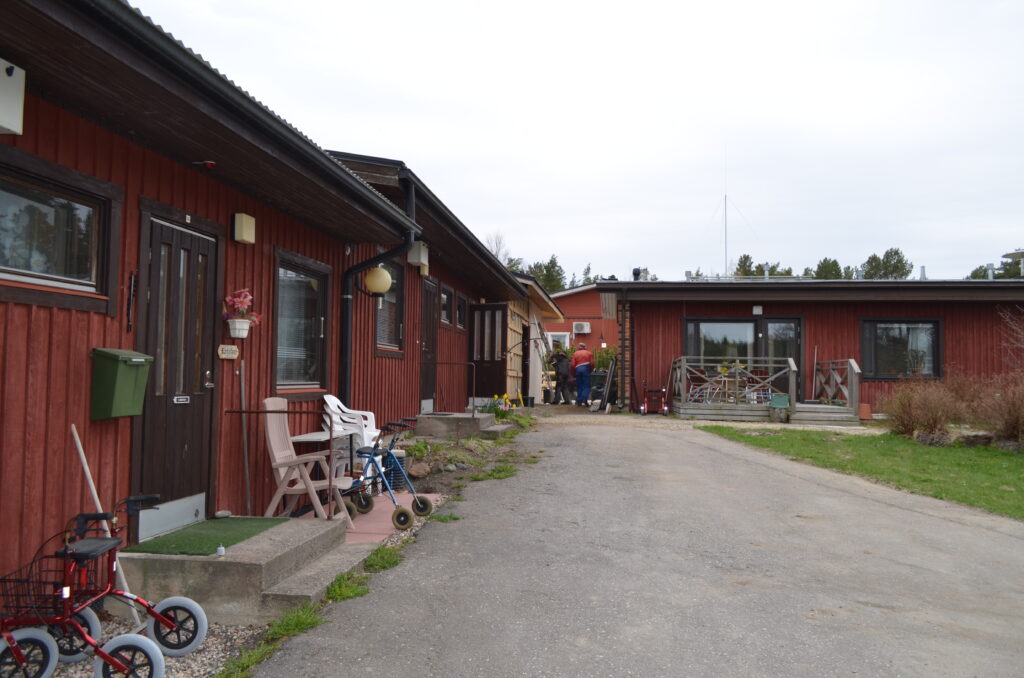 En asfalterad gångväg löper mellan envåningshus i trä med bruna och röda fasader. Rullatorer står uppställda vid dörröppningarna. En grupp människor står nära ett av husen i bakgrunden. Himlen är molnig.