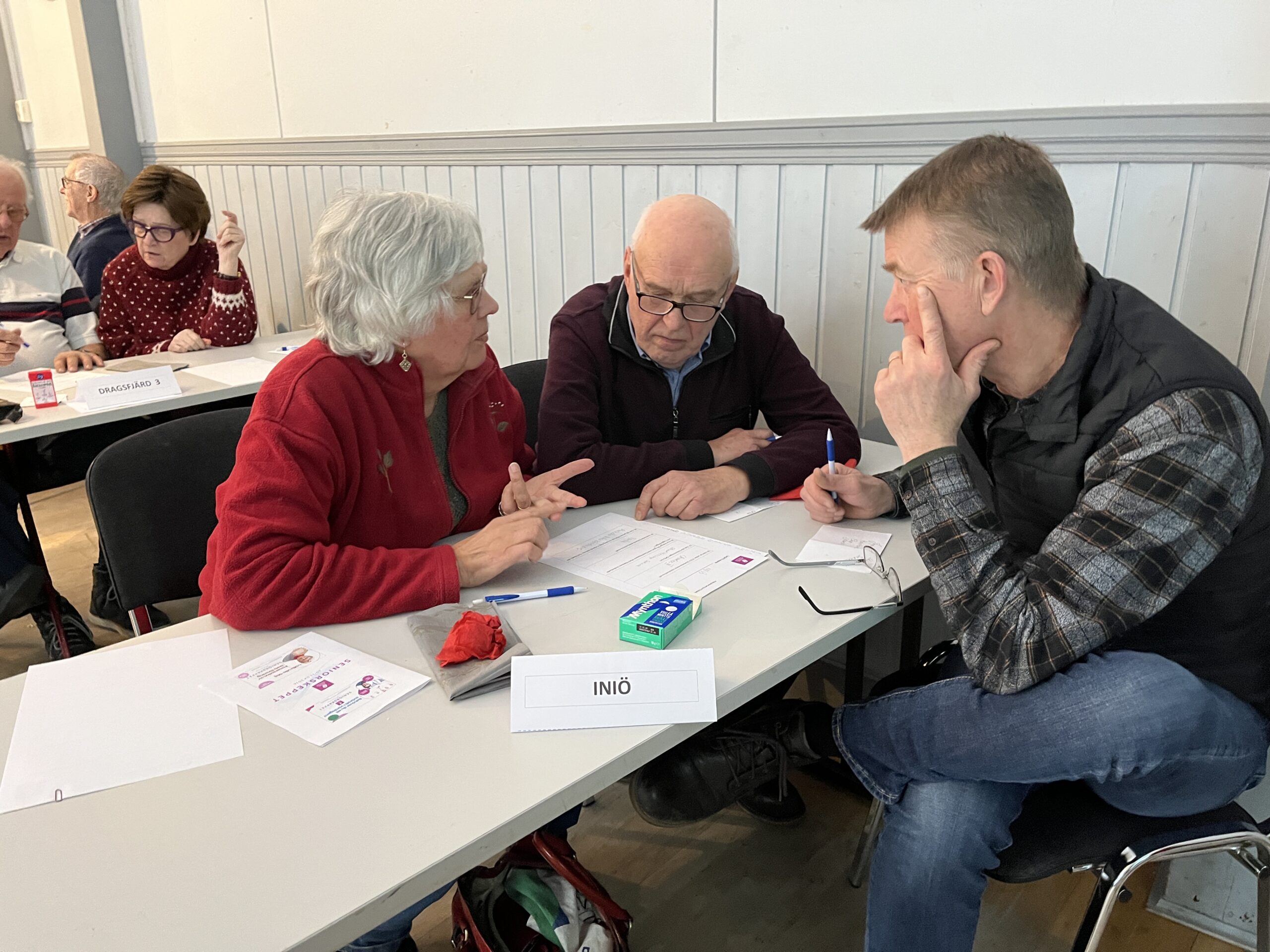 Tre vuxna sitter vid ett bord och diskuterar, med blicken riktad mot papperen framför sig. På bordet står en skylt med texten ”INIÖ”. I bakgrunden syns andra grupper som också arbetar tillsammans.