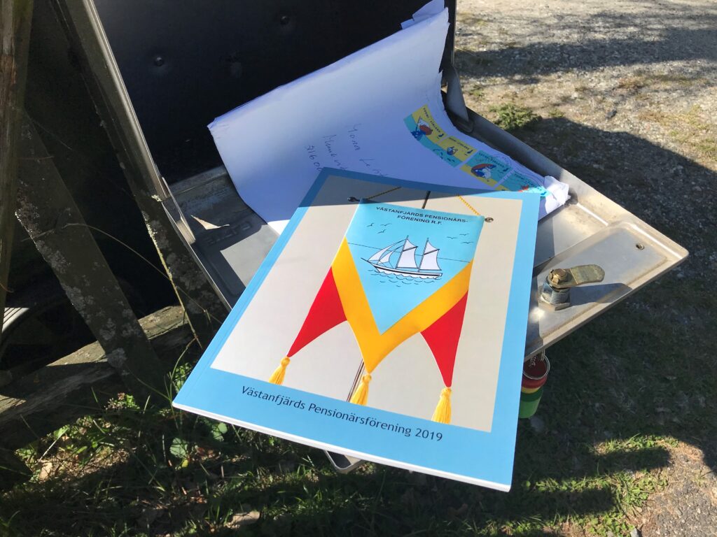 En ljusblå broschyr med titeln ”Västanfjärds Pensionärsförening 2019”, med en illustration av en segelbåt och flaggor, ligger ovanpå en hög med post i en öppen metallbrevlåda utomhus.