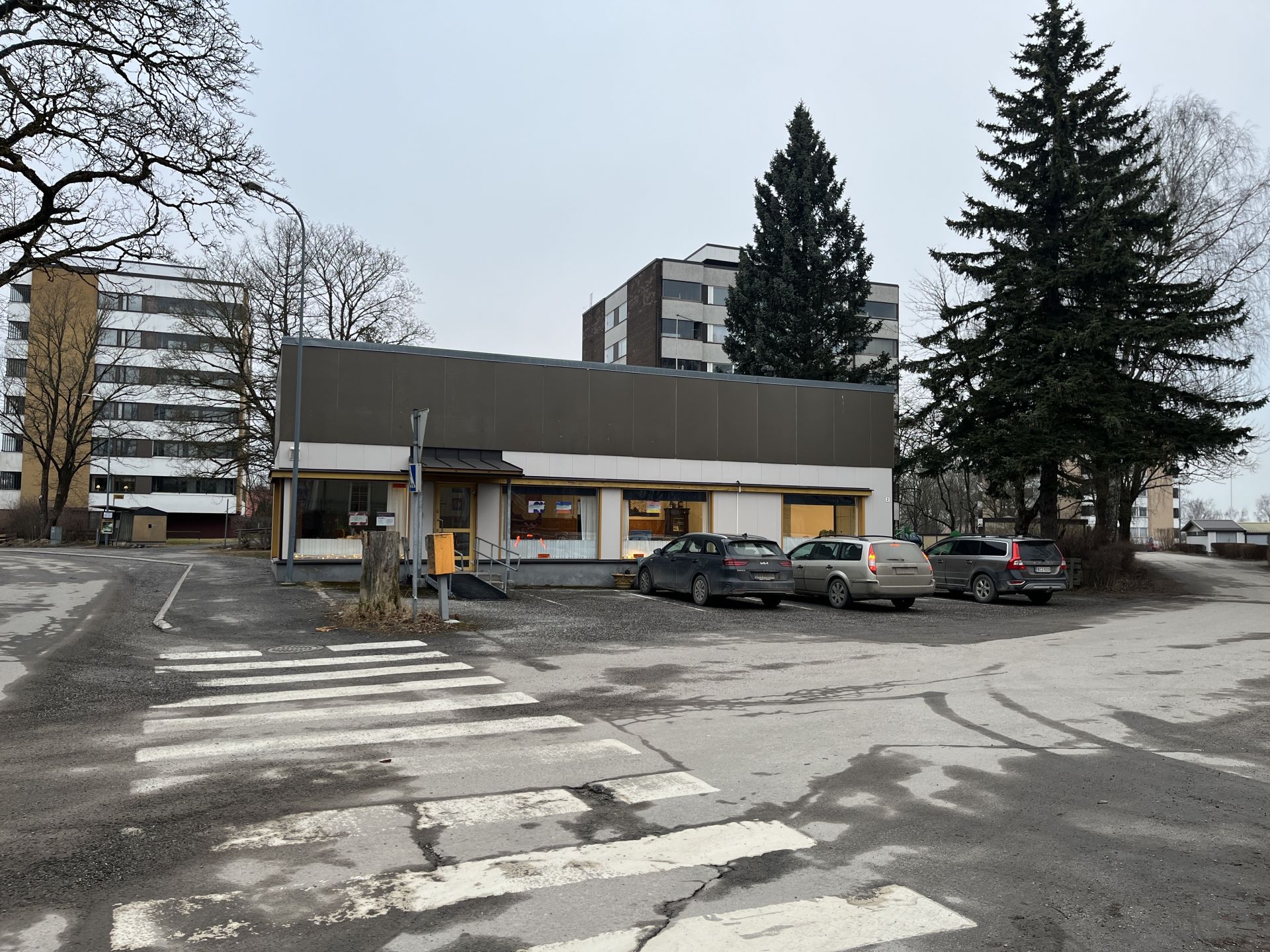 En liten, envåningsbyggnad med stora fönster ligger framför tre parkerade bilar. I bakgrunden syns flerbostadshus och höga träd. I förgrunden syns ett övergångsställe och en vägskylt.