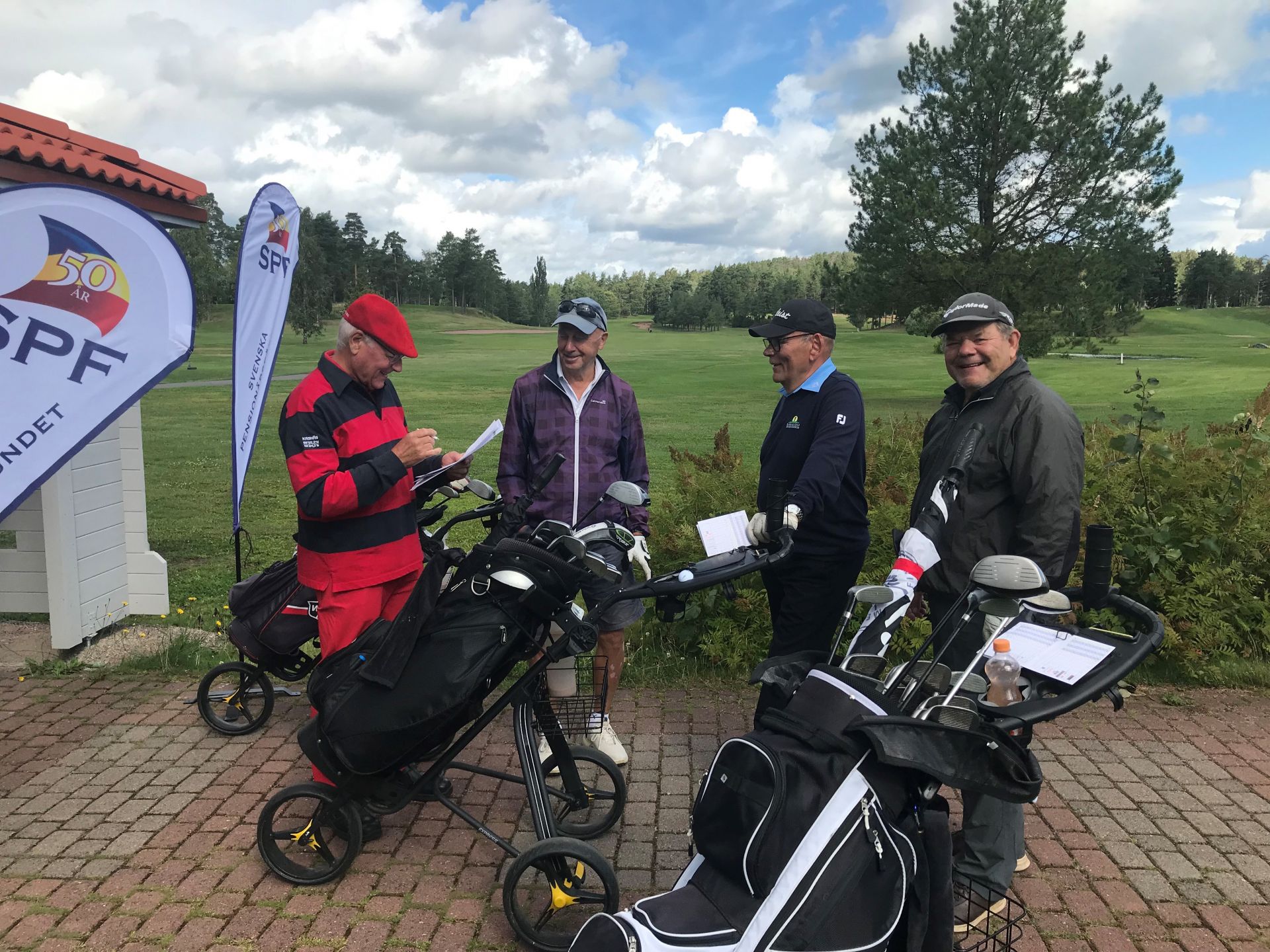 Fyra män med golfväskor står på en tegelgång nära en golfbana och pratar och ler. Till vänster syns en banderoll och en byggnad, och i bakgrunden syns gröna fairways med träd under en delvis molnig himmel.