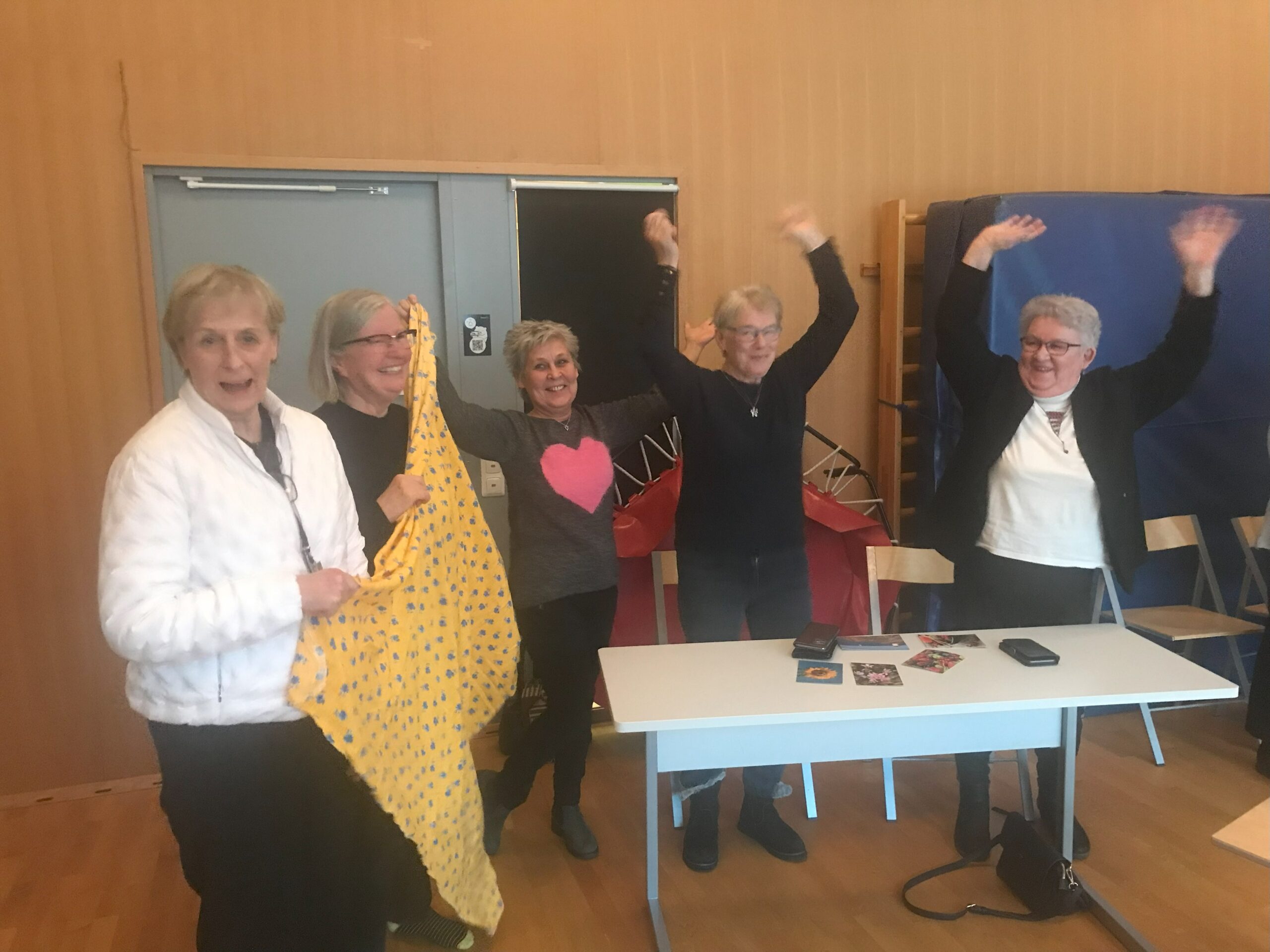 Fem äldre kvinnor står tillsammans inomhus, några med armarna uppsträckta, och ler. En av dem håller i ett gult mönstrat tygstycke. De står bredvid ett bord med några små föremål och en telefon på. I bakgrunden syns hopfällbara stolar och mattor.