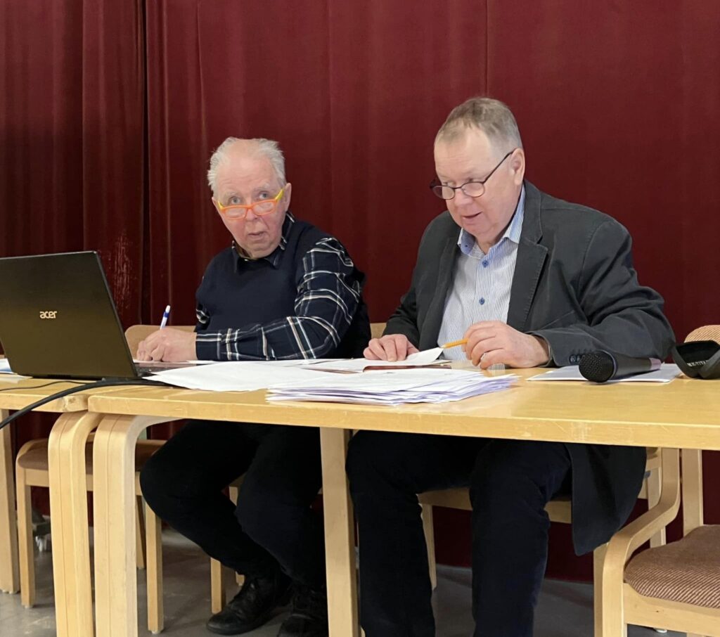Två äldre män sitter vid ett bord täckt av papper och en bärbar dator, framför en röd gardin. Den ene mannen skriver med en penna medan den andre läser dokument. En mikrofon ligger på en stol i närheten.