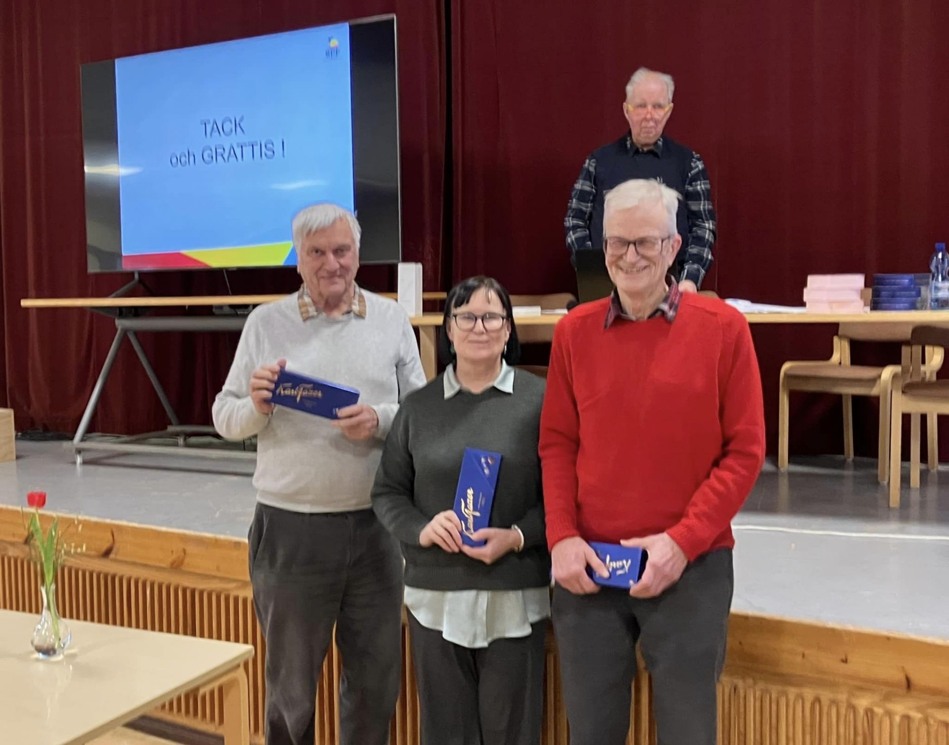 Tre personer står framför en scen med blå kuvert i händerna och poserar för ett foto. En man står bakom dem på scenen. På en skärm i bakgrunden syns orden ”TACK och GRATTIS!”, och på ett bord står olika föremål och stolar.