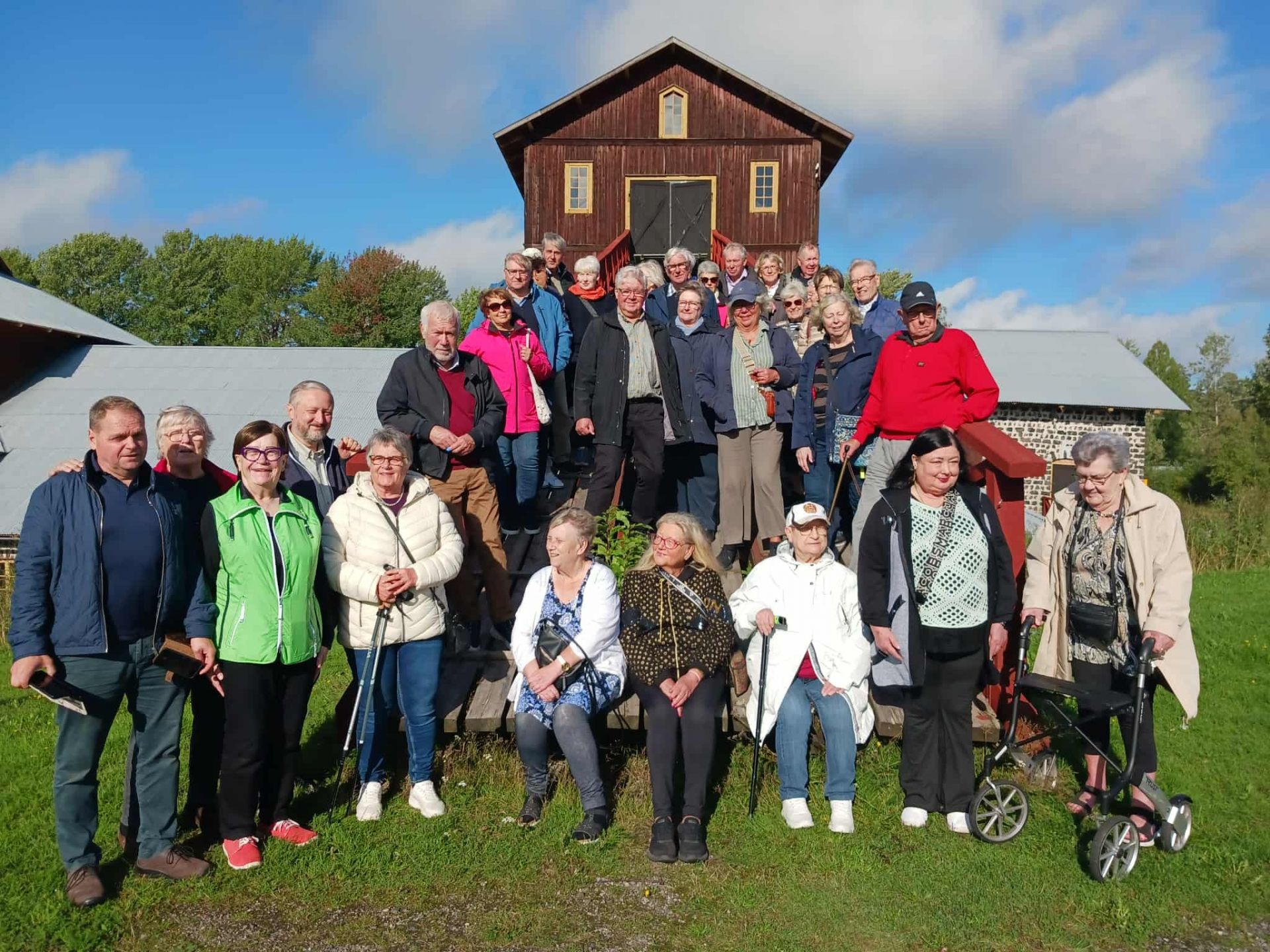 En grupp på cirka 30 äldre vuxna poserar tillsammans utomhus framför en trälada en solig dag. Vissa står på trappsteg, medan andra sitter eller står framför ladan, med grönt gräs och blå himmel i bakgrunden.