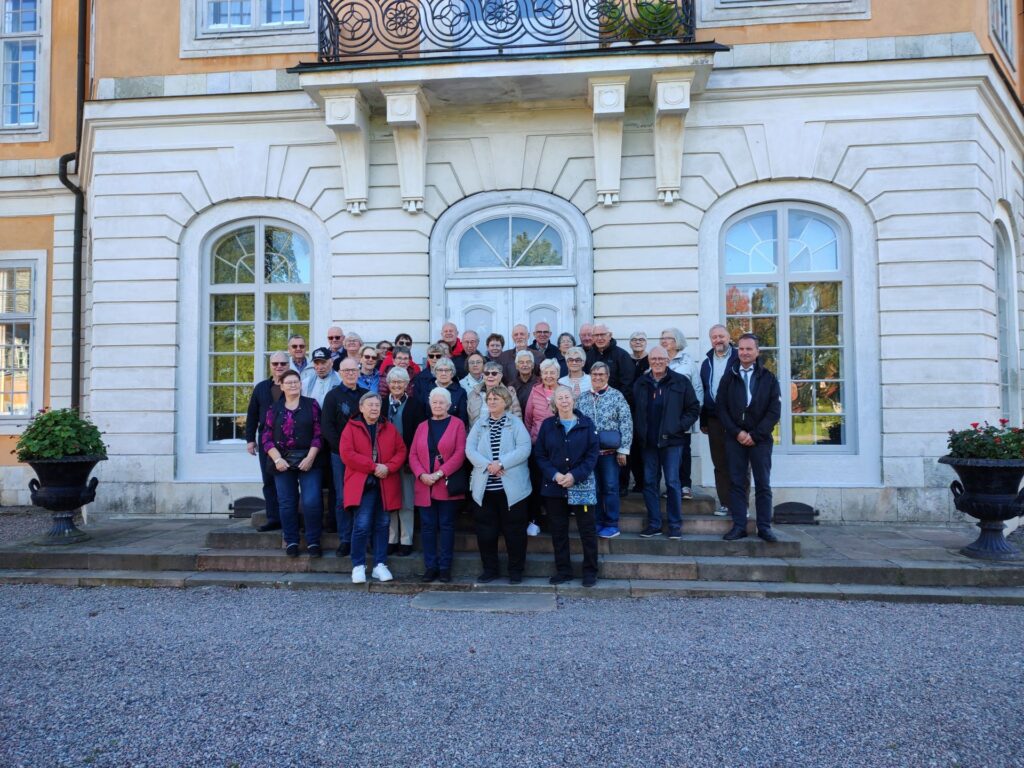 En grupp på omkring 30 personer står på trappan framför en stor byggnad med välvda fönster och vita stenväggar och poserar för en gruppbild en klar dag.