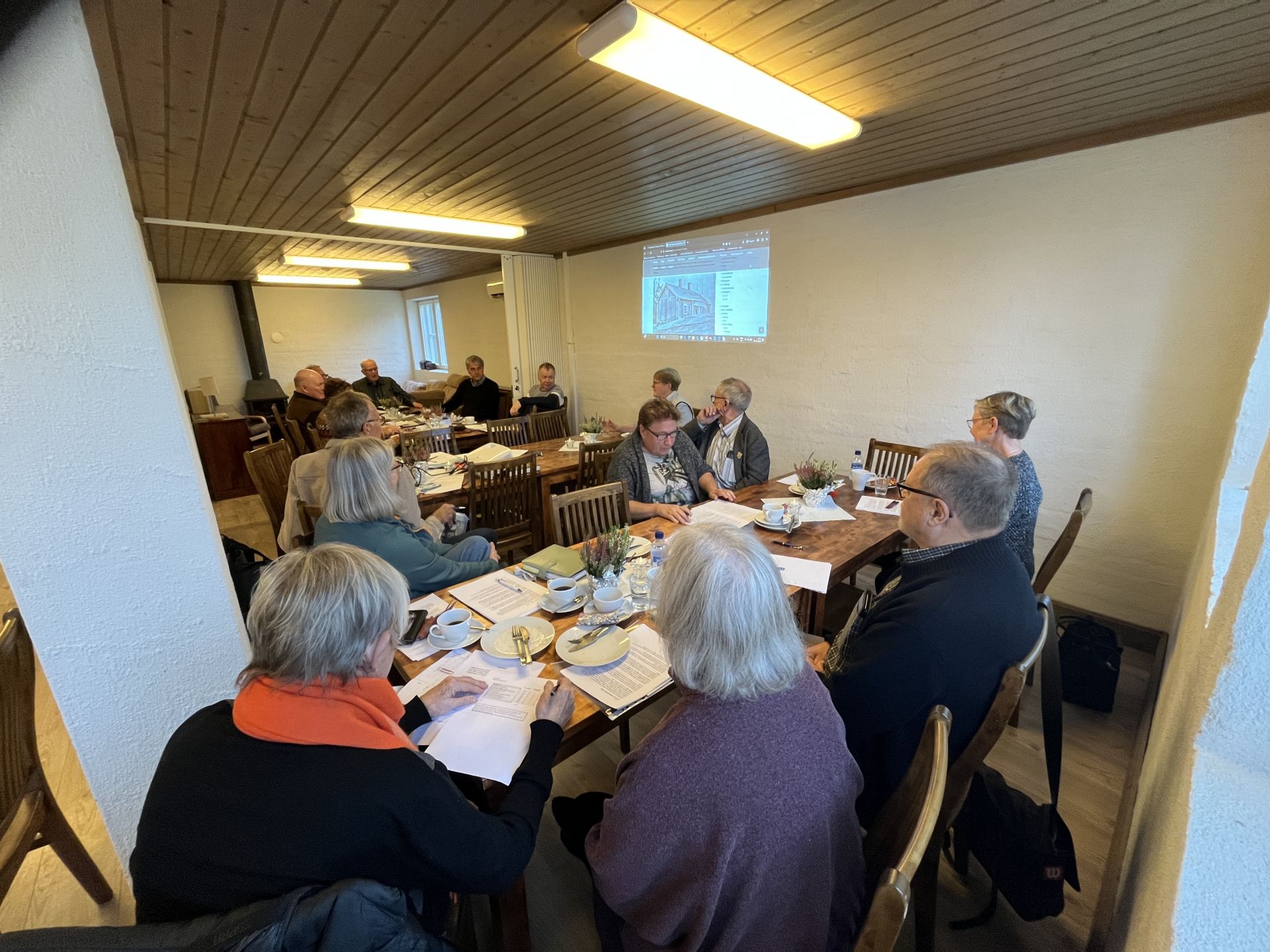 En grupp människor sitter runt bord i ett mötesrum med papper, drycker och snacks framför sig. En presentation projiceras på väggen i bakgrunden. Rummet har trämöbler och ett trätak.