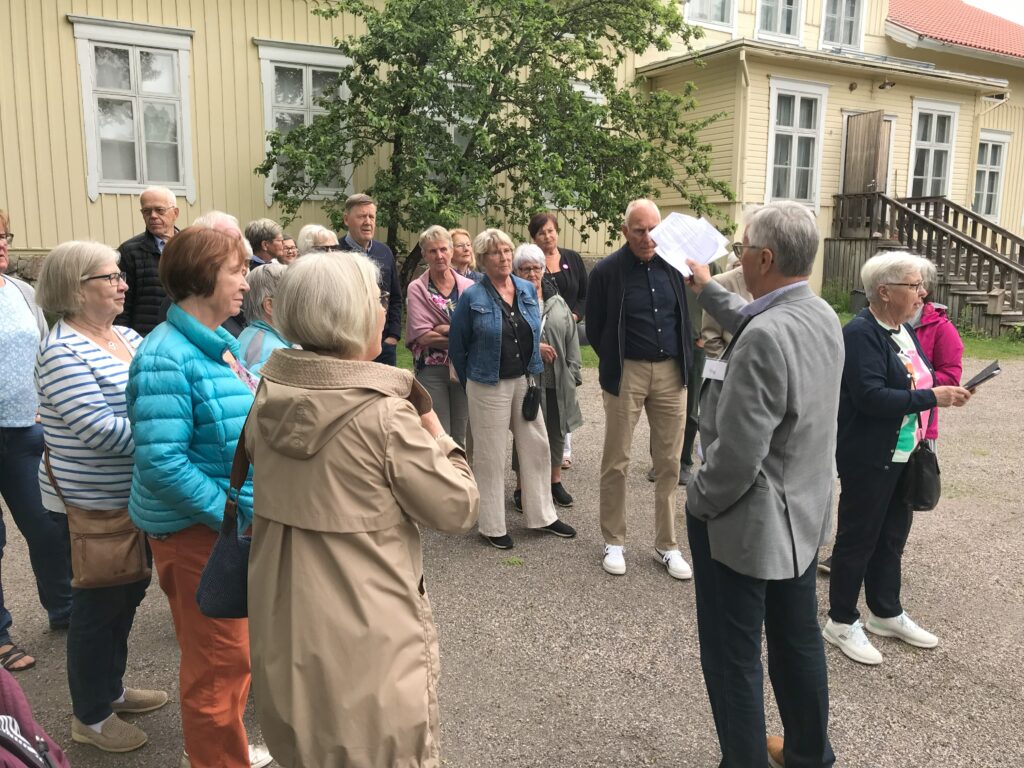 En grupp vuxna står utanför en gul byggnad och lyssnar på en person som håller i några papper och som verkar hålla en rundvisning eller ge en förklaring. Scenen utspelar sig utomhus, och i bakgrunden syns ett träd och en trappa.