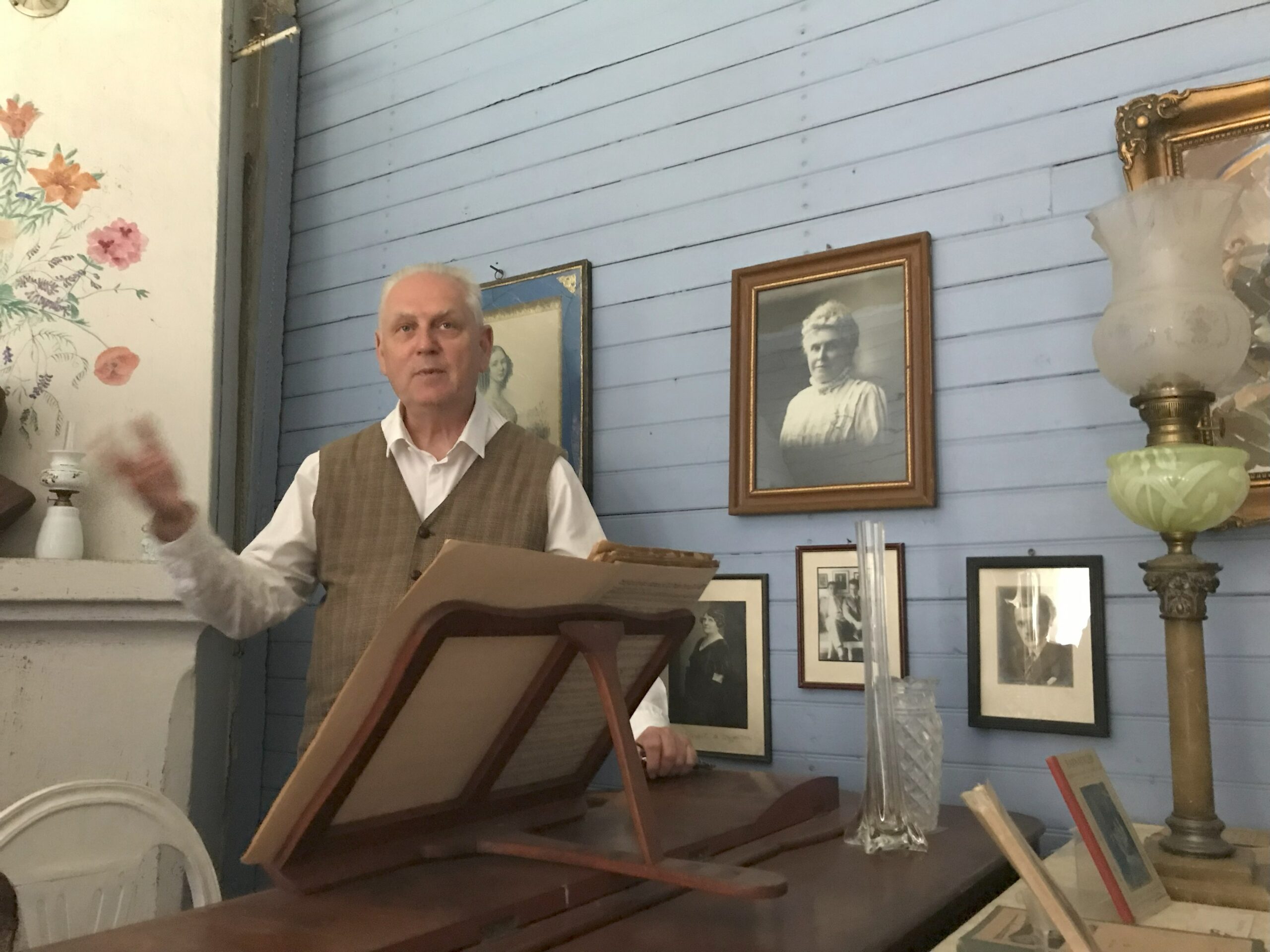 En äldre man står bakom ett notställ av trä i ett rum i retrostil med ljusblå träväggar, dekorerat med inramade svartvita porträtt och en antik lampa bredvid honom.
