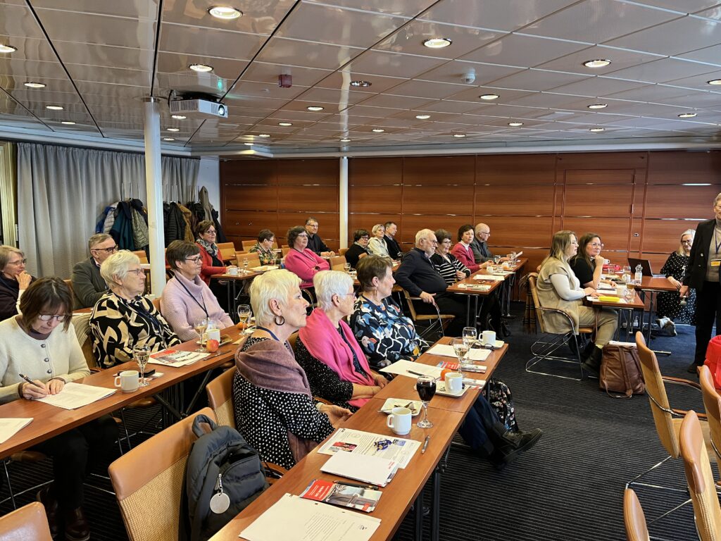 En grupp vuxna sitter vid bord i ett konferens- eller mötesrum, vända framåt. Vissa skriver på papper, medan andra lyssnar. På borden står kaffekoppar och papper. Rummet har träpaneler och ett kaklat tak.
