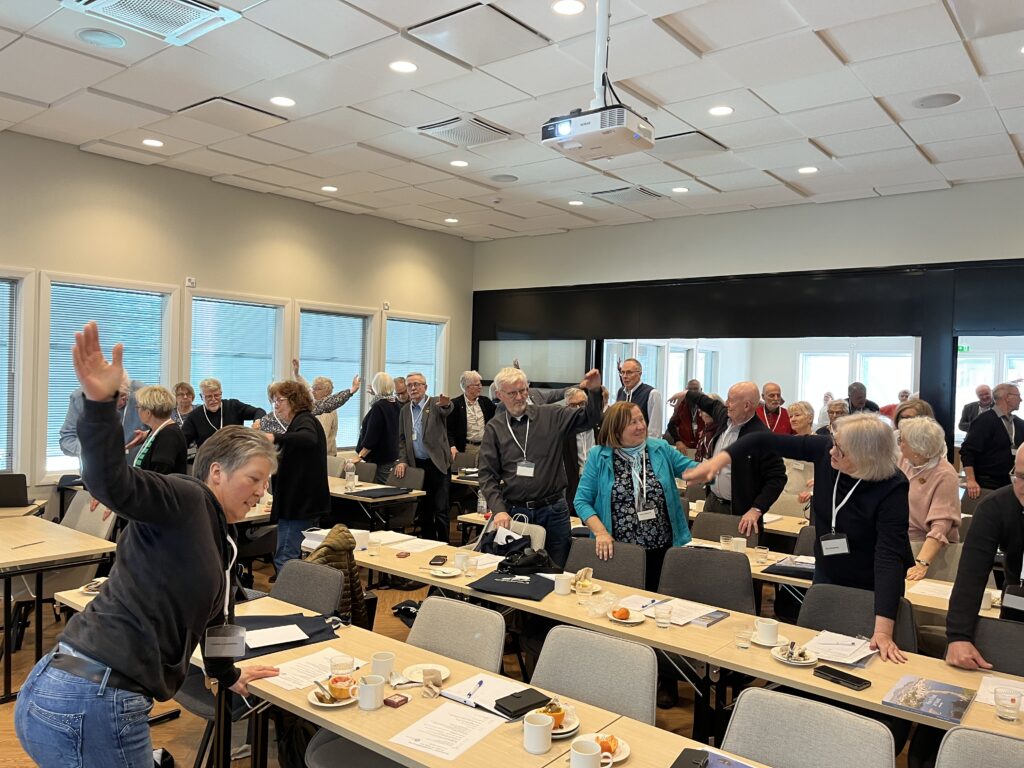 En grupp vuxna står vid borden i ett konferensrum och sträcker upp ena armen, som om de deltog i en gruppaktivitet eller övning. Rummet har stora fönster, beige väggar och en projektor monterad i taket.